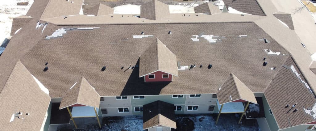 Wind damage on the roof of an apartment building. Shingles are blown off in sections revealing the synthetic underlayment.